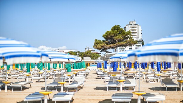 A beach with rows of sun loungers and umbrellas, a clear sky, and a building in the background. - Hotel Bella Venezia Mare (Lignano Sabbiadoro)