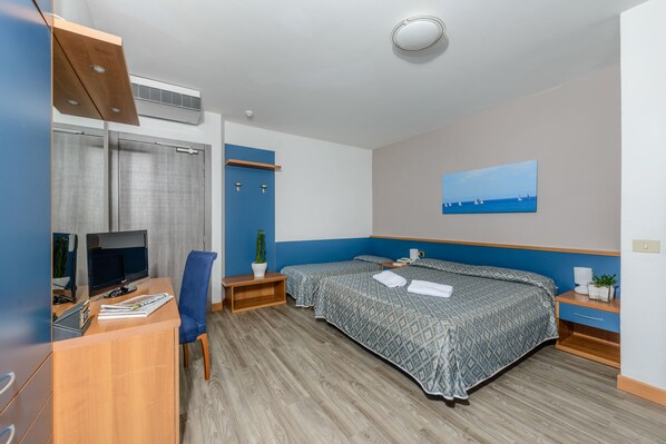 Habitación de hotel con escritorio, dos camas, televisor y un cuadro de veleros en la pared.