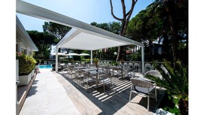 An outdoor seating area with tables and chairs under a white canopy, surrounded by greenery and trees. - Hotel Bella Venezia Mare (Lignano Sabbiadoro)