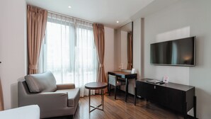 A modern hotel room with a sofa, a small round table, a desk, and a flat-screen TV mounted on the wall.