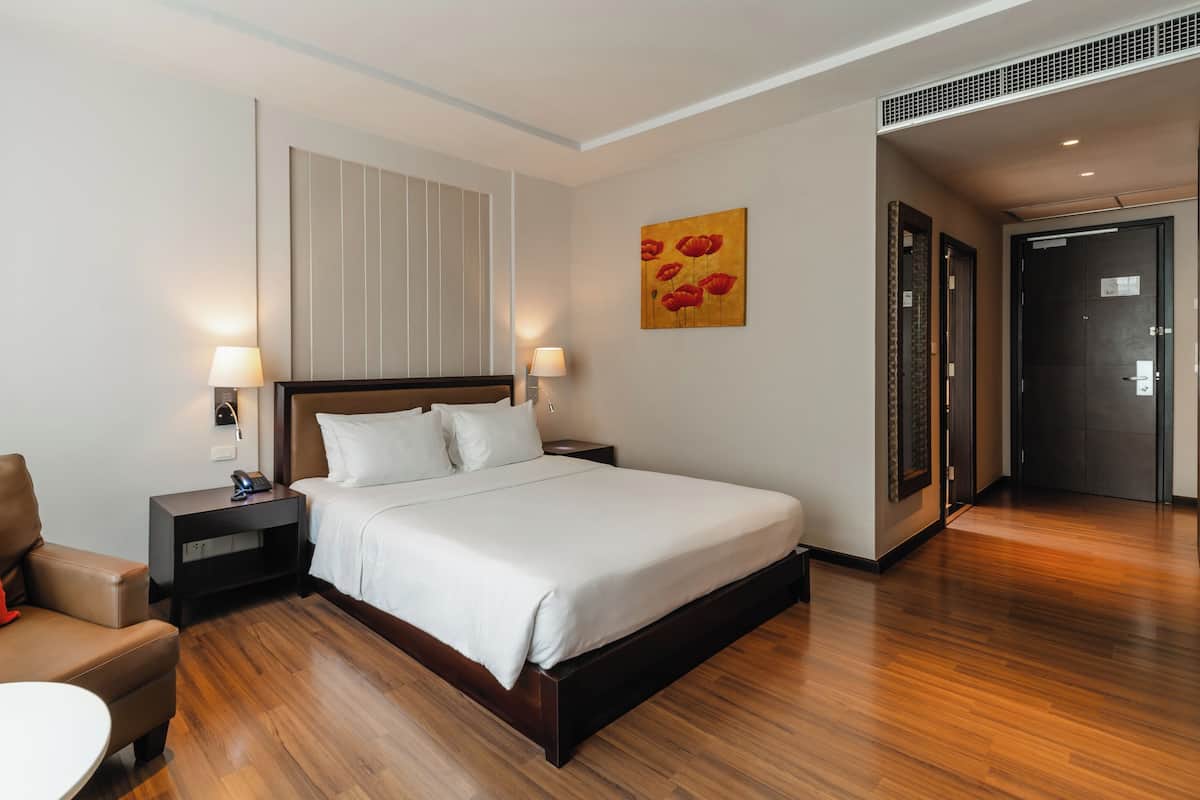 Een hotelkamer met een bed, nachtkastje met lamp, een stoel en een schilderij aan de muur.