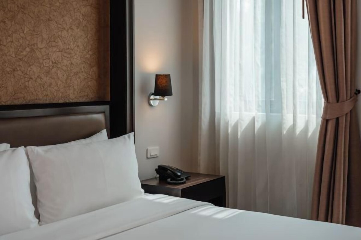 A hotel room with a bed, a nightstand, a lamp, and sheer curtains.