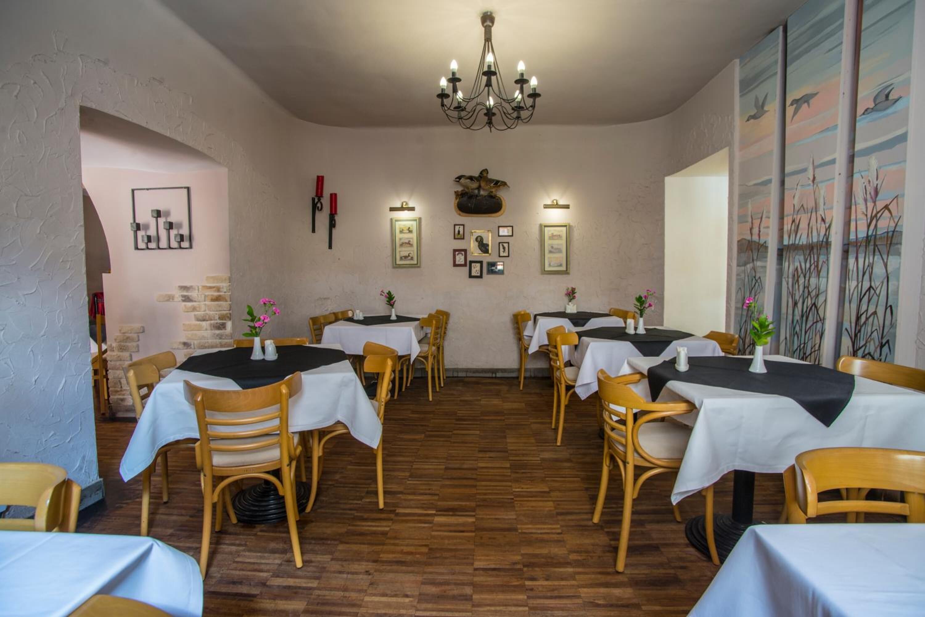 A restaurant with wooden chairs and tables, a chandelier, framed pictures on the wall, and a brick wall.