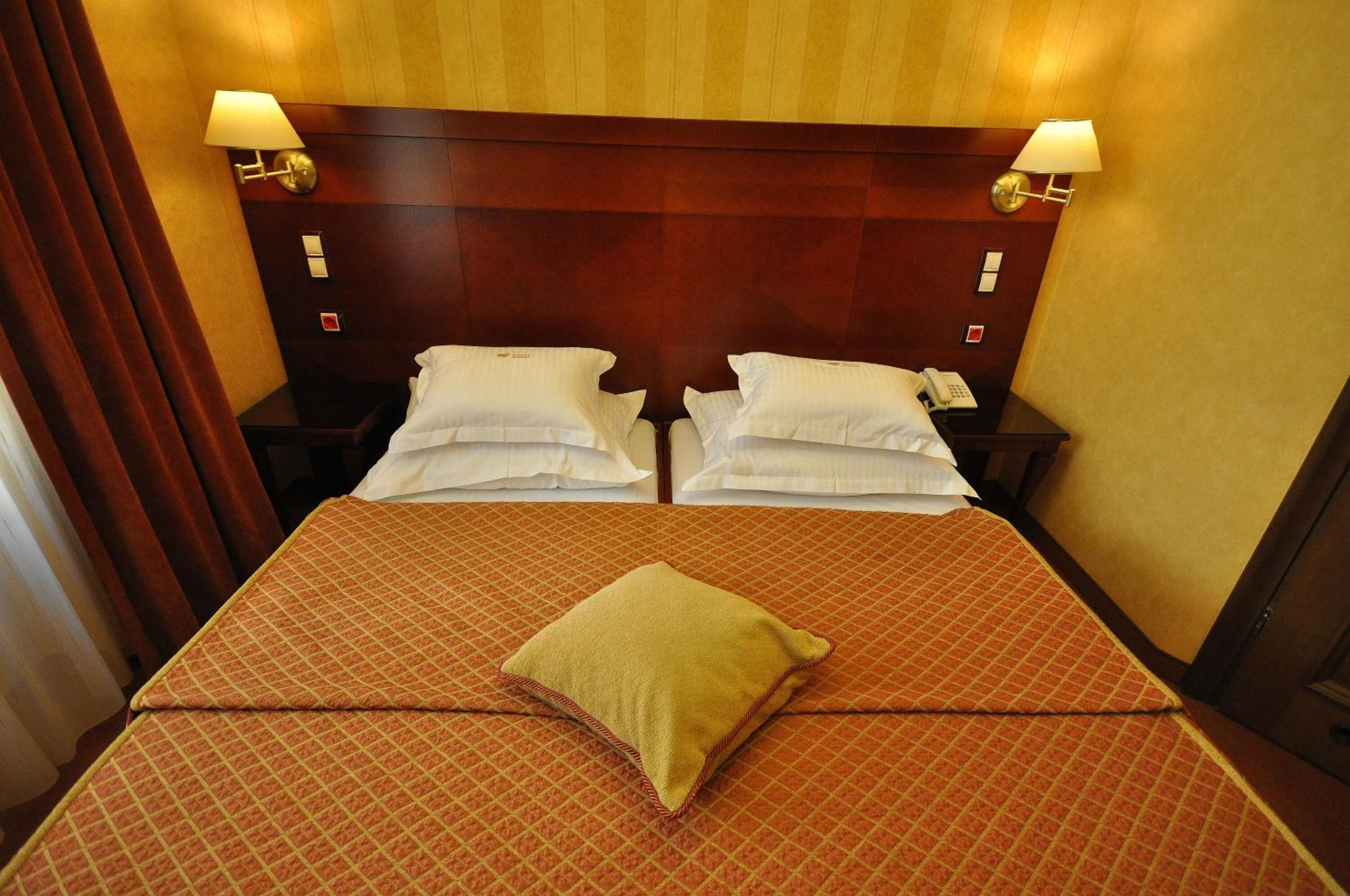 Uma cama de casal com colcha estampada, duas mesas de cabeceira com abajures e uma cabeceira de madeira.