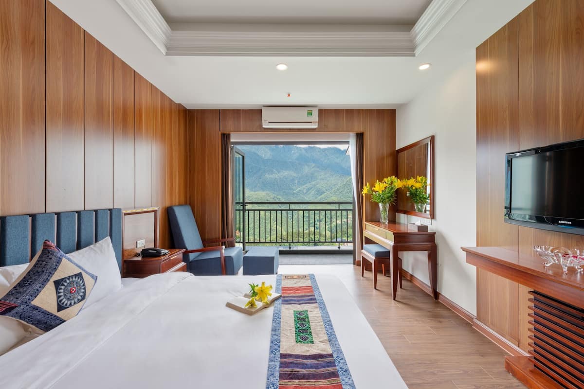 A hotel room with a large bed, a desk, a television, and a balcony with a mountain view.