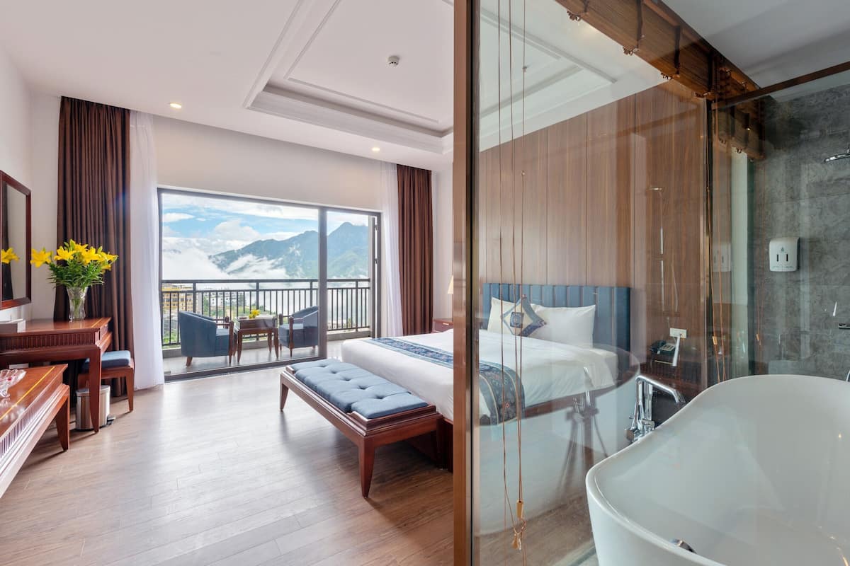A modern hotel room with a large bed, a glass-enclosed bathroom, a balcony with a mountain view, and a dining area with wooden furniture.