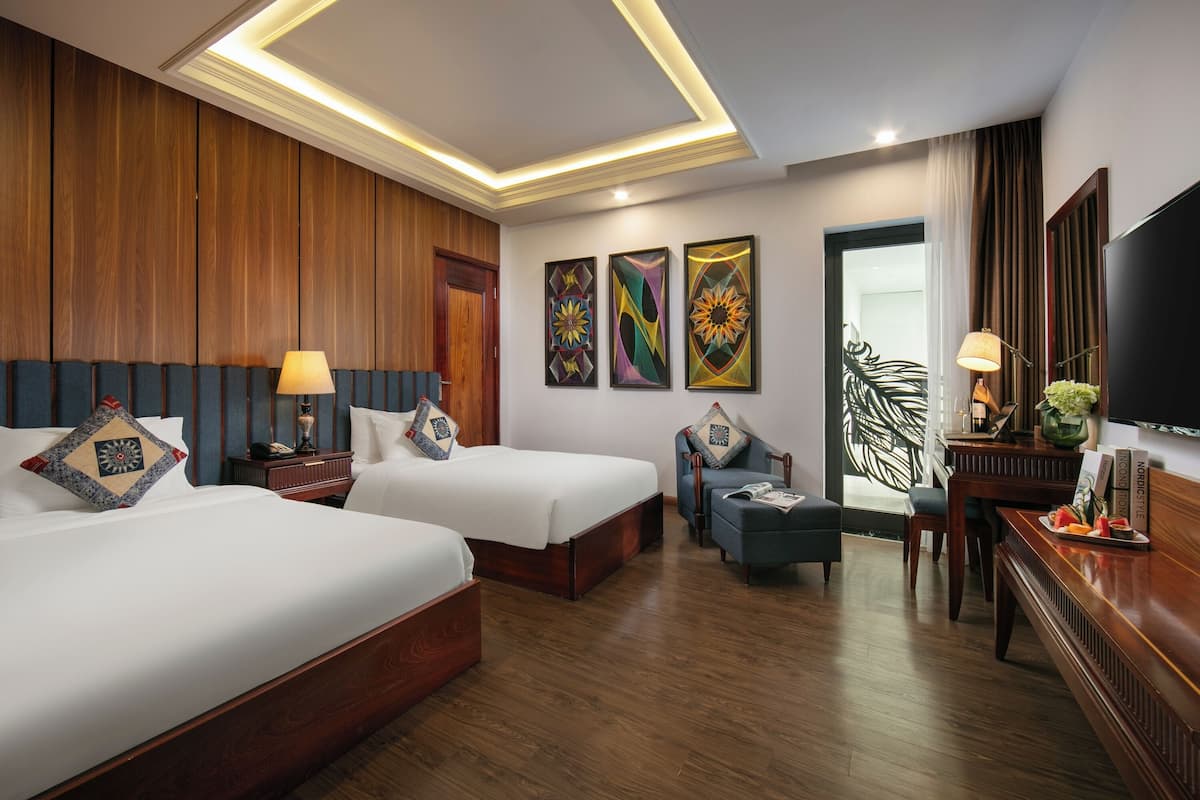 A hotel room with two beds, wooden paneling, a TV, artwork, and a desk with a lamp.