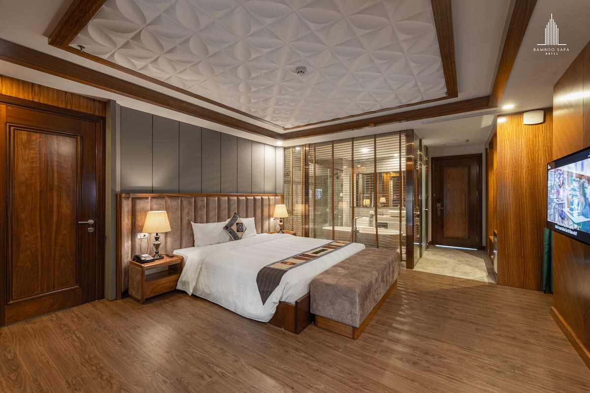 A spacious hotel room with a large bed, wooden flooring, a modern ceiling design, and a television on the wall.