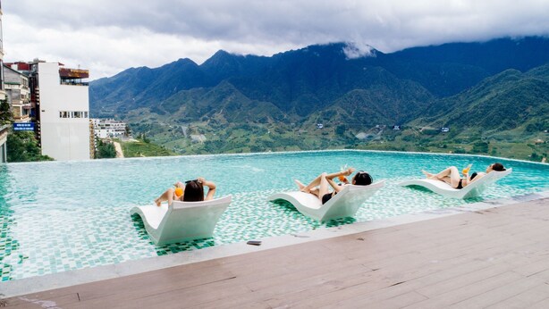 Piscina com três espreguiçadeiras, piso ladrilhado e montanhas ao fundo.