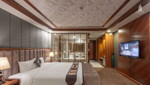 Quarto de hotel com uma cama grande, mesas de cabeceira, uma televisão e um banheiro visível através de um painel de vidro.