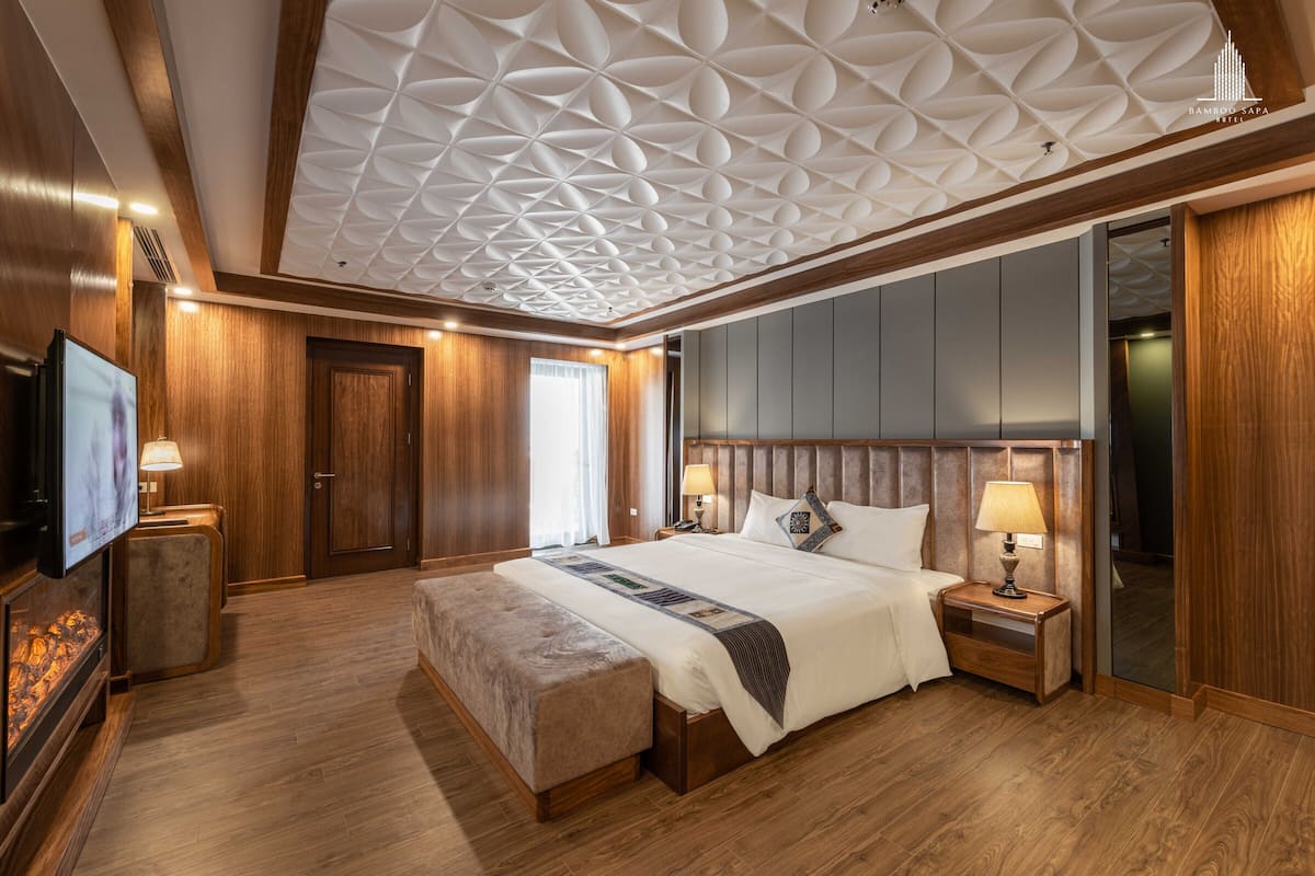 A hotel room with a wooden ceiling, a large bed with a headboard, bedside tables with lamps, a television, and a wooden floor.