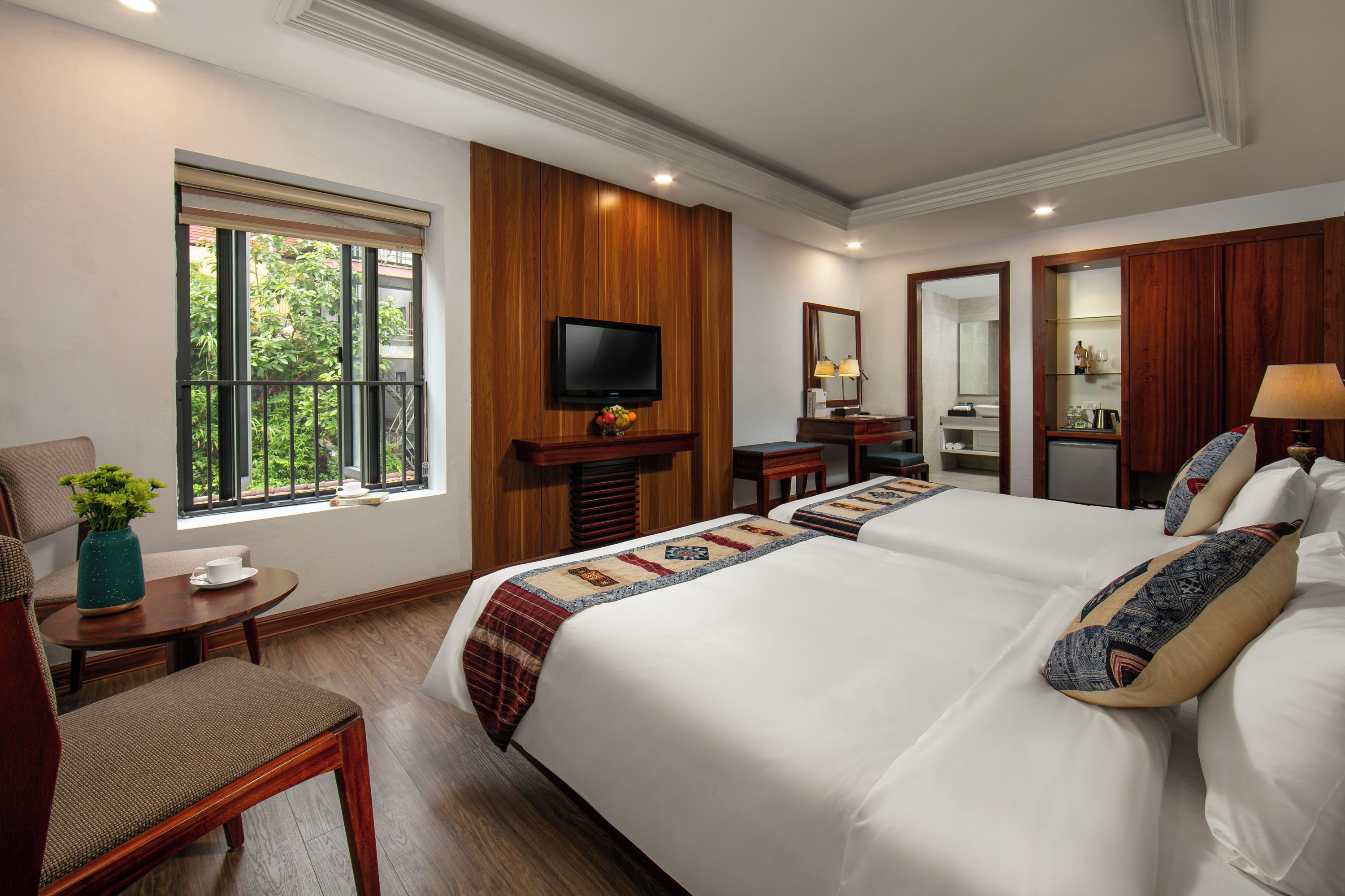 Habitación de hotel con una cama grande, televisión, escritorio con silla, una mesita con un florero y ventana con vistas a la vegetación.