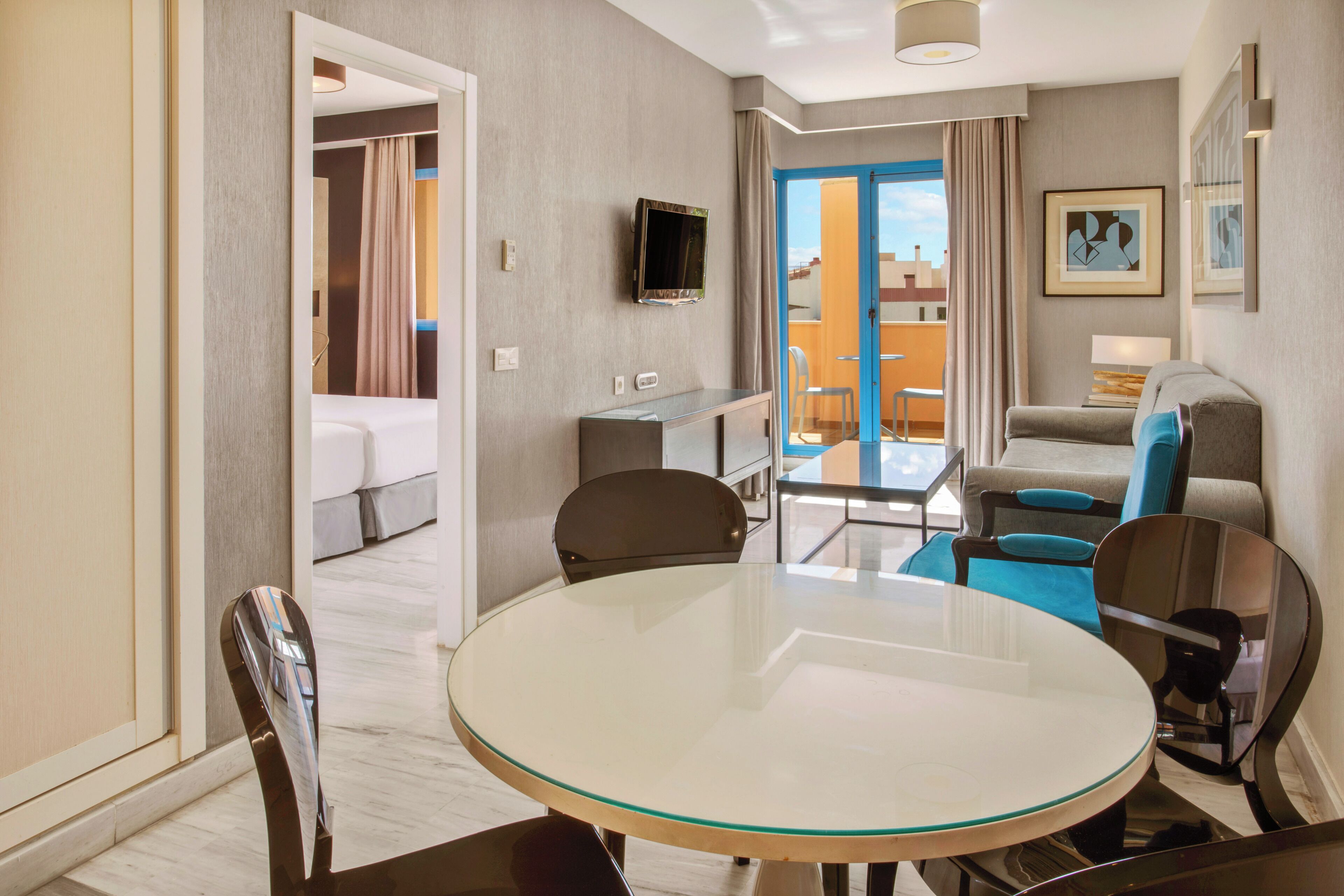A hotel room with a round glass table, a bed, a TV, a balcony with a view, and a sofa.
