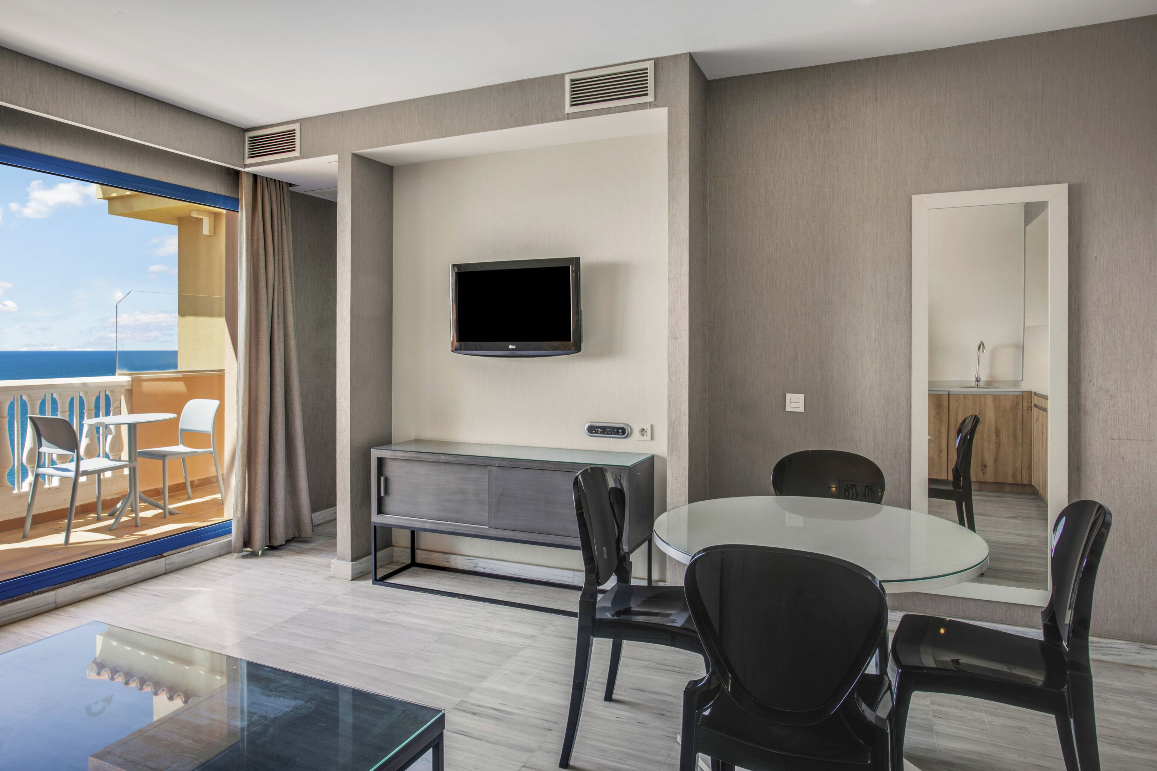 A modern hotel room with a balcony, a flat-screen TV, a glass dining table, and a compact kitchenette.