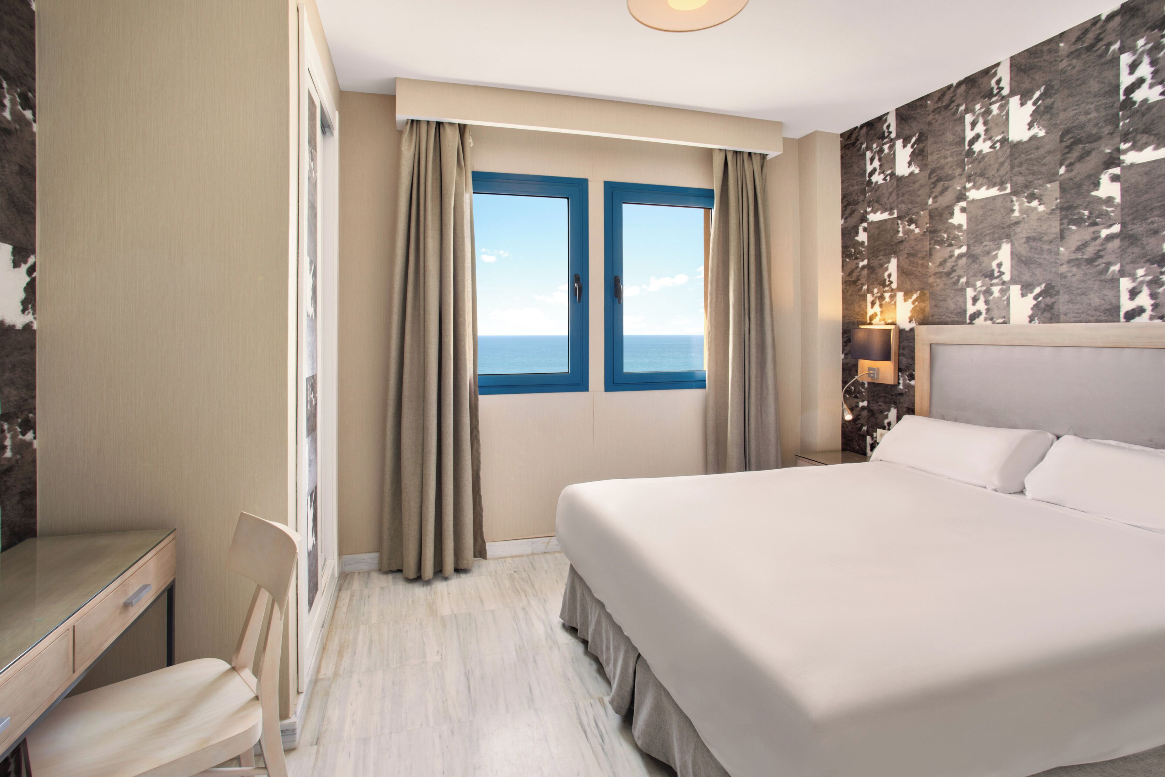 Een hotelkamer met een bed, een bureau, een stoel, een raam met gordijnen en een wand met een patroon.