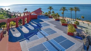 Ein Tennisplatz mit rotem Bogen, Palmen und Blick auf das Meer.