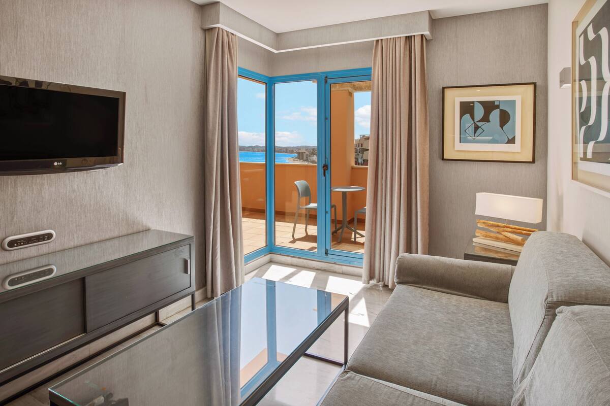 A hotel room with a flat-screen TV, a glass coffee table, a sofa, a dining area with a table and chairs, and a view of the sea through a glass door.