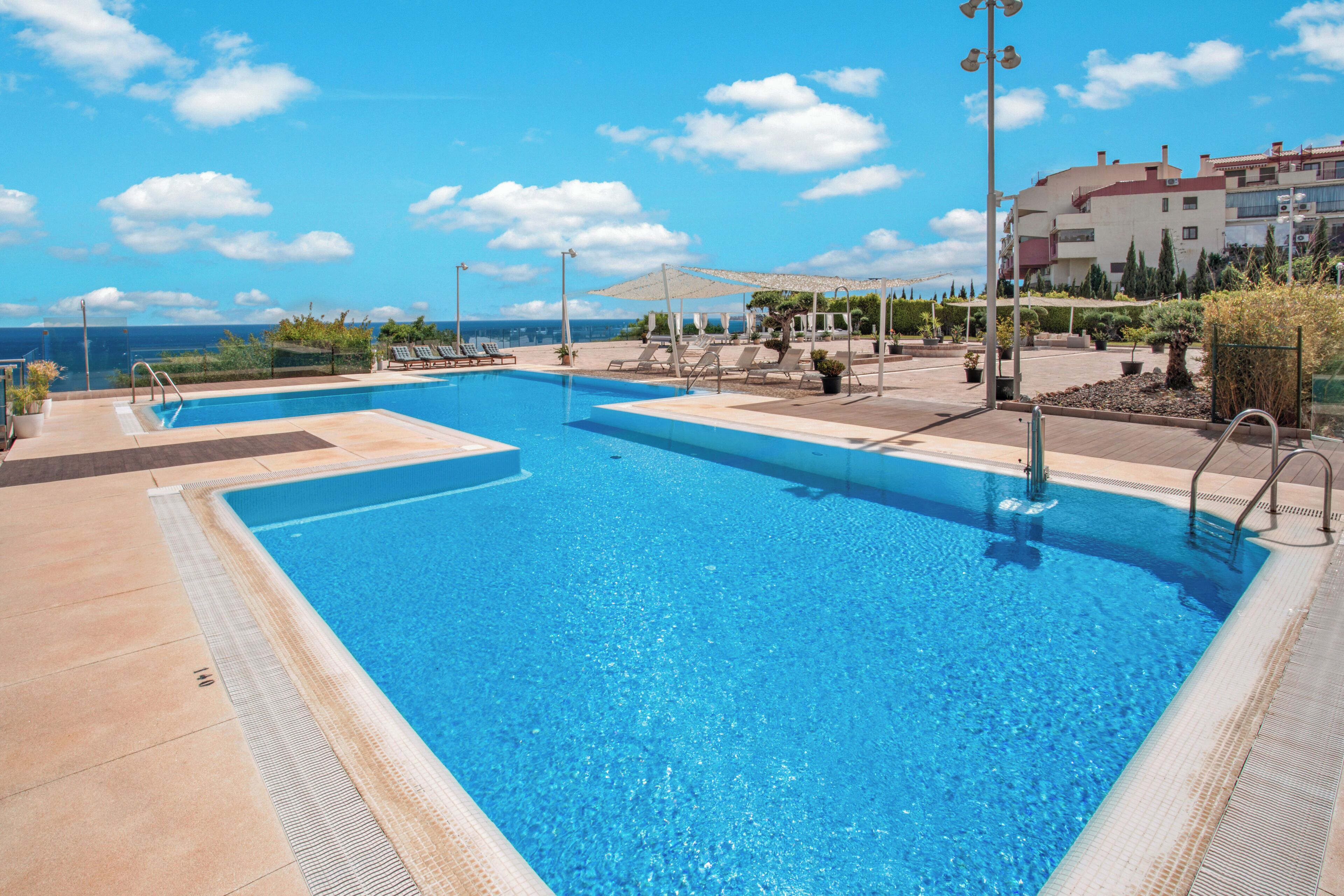 Una piscina con agua azul cristalina, rodeada de sillas de descanso y sombrillas, con vista al mar y edificios al fondo.