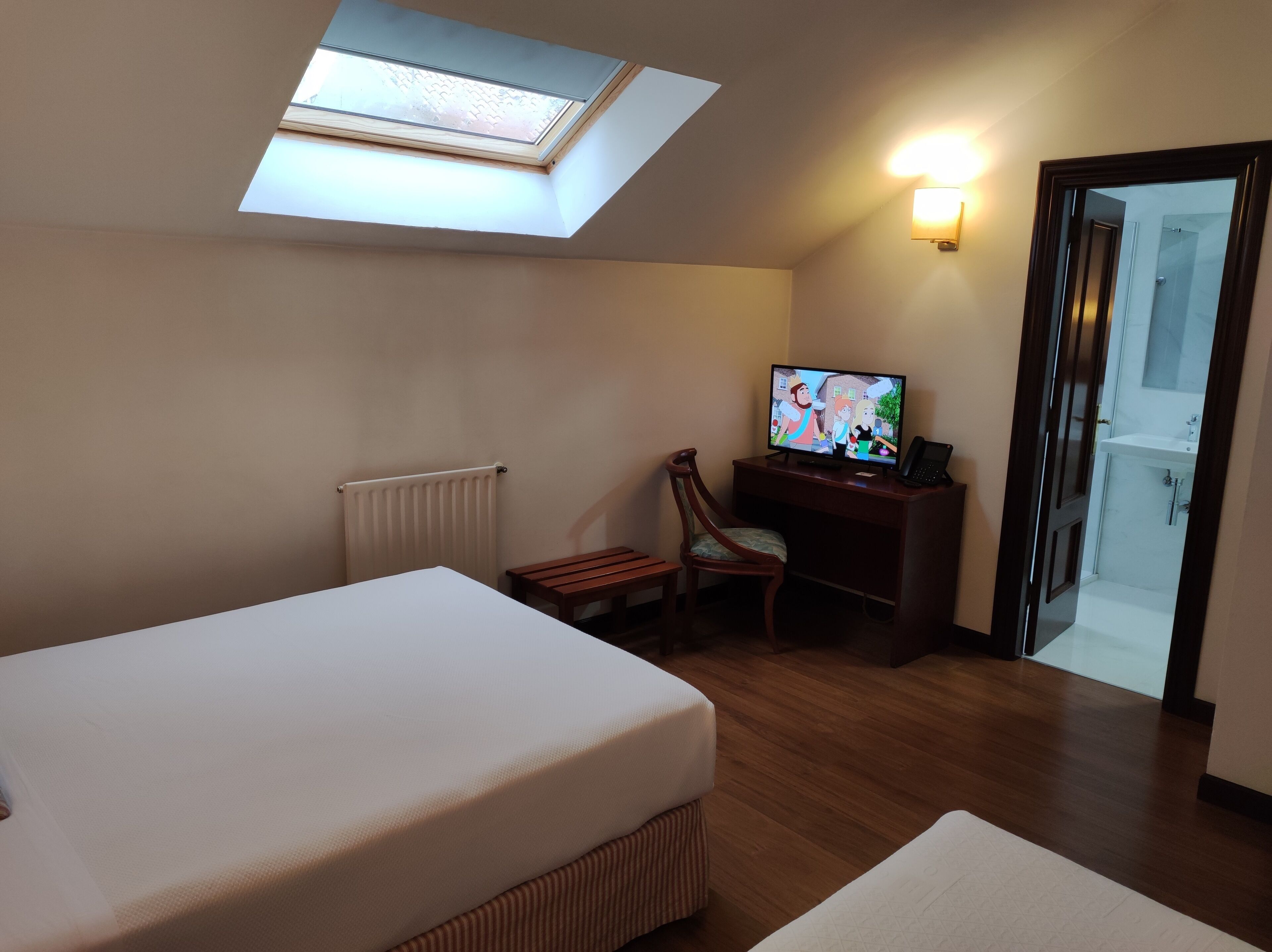 Ein Hotelzimmer mit Oberlicht, einem Bett, einem Schreibtisch mit Computer und einem durch eine offene Tür einsehbaren Badezimmer.