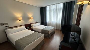 Habitación de hotel con dos camas individuales, un escritorio con silla, televisión y ventana con cortinas.