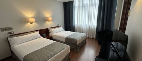 A hotel room with two single beds, a desk with a chair, a TV, and a window with curtains.