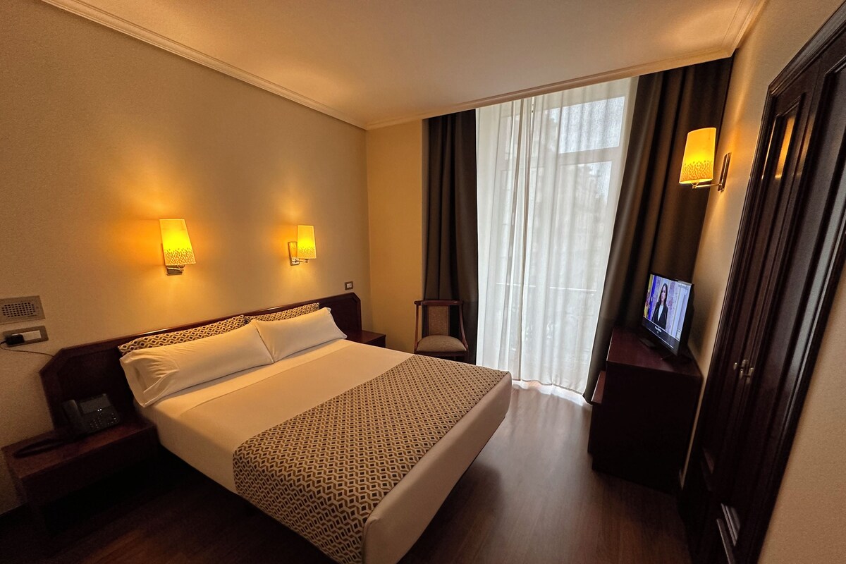 A hotel room with a large bed, two bedside lamps, a TV, and a view of the outdoors through a window with curtains.