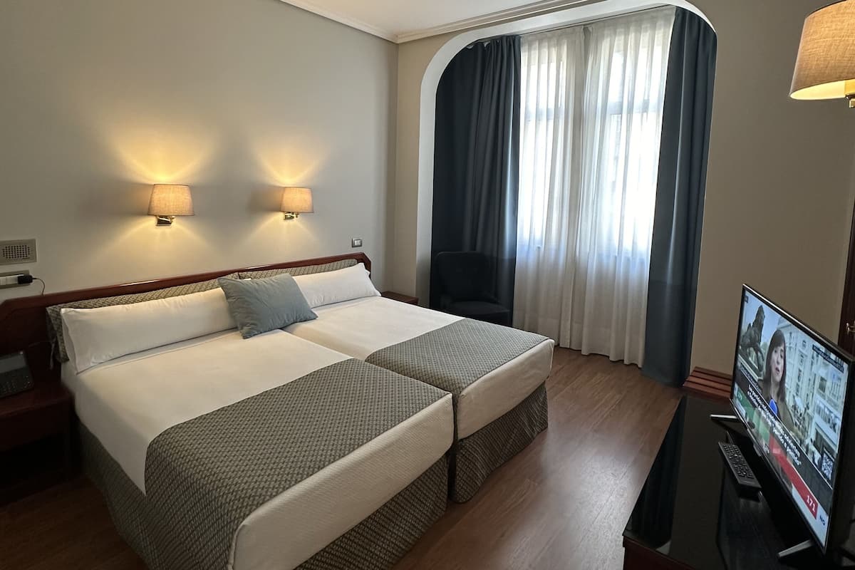 A hotel room with a double bed, two bedside lamps, a TV, and a window with curtains.