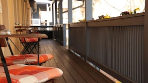 Un espace extérieur abrité, avec des tables et des chaises en bois, une rambarde et une vue sur la rue.