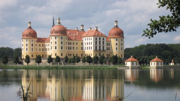 Un grand bâtiment historique, doté de plusieurs toits en forme de dôme et d’une façade symétrique, situé au bord d’un lac paisible dont les eaux reflètent le ciel.