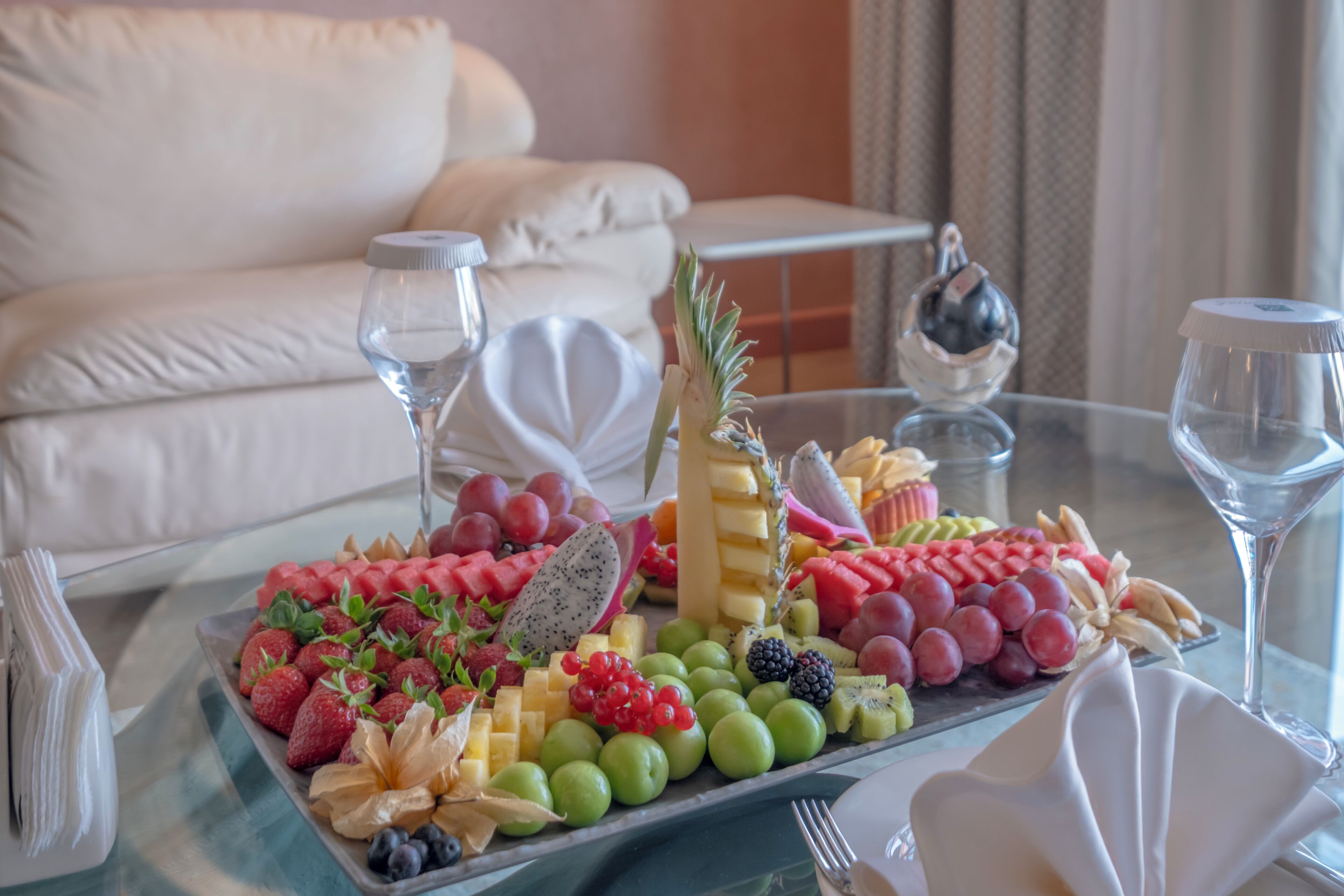 Una bandeja de frutas sobre una mesa de vidrio, con un sofá blanco al fondo.