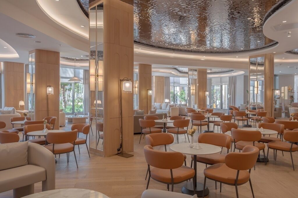 A spacious hotel lobby with round tables and orange chairs, large wooden pillars, and a high ceiling with a reflective surface.