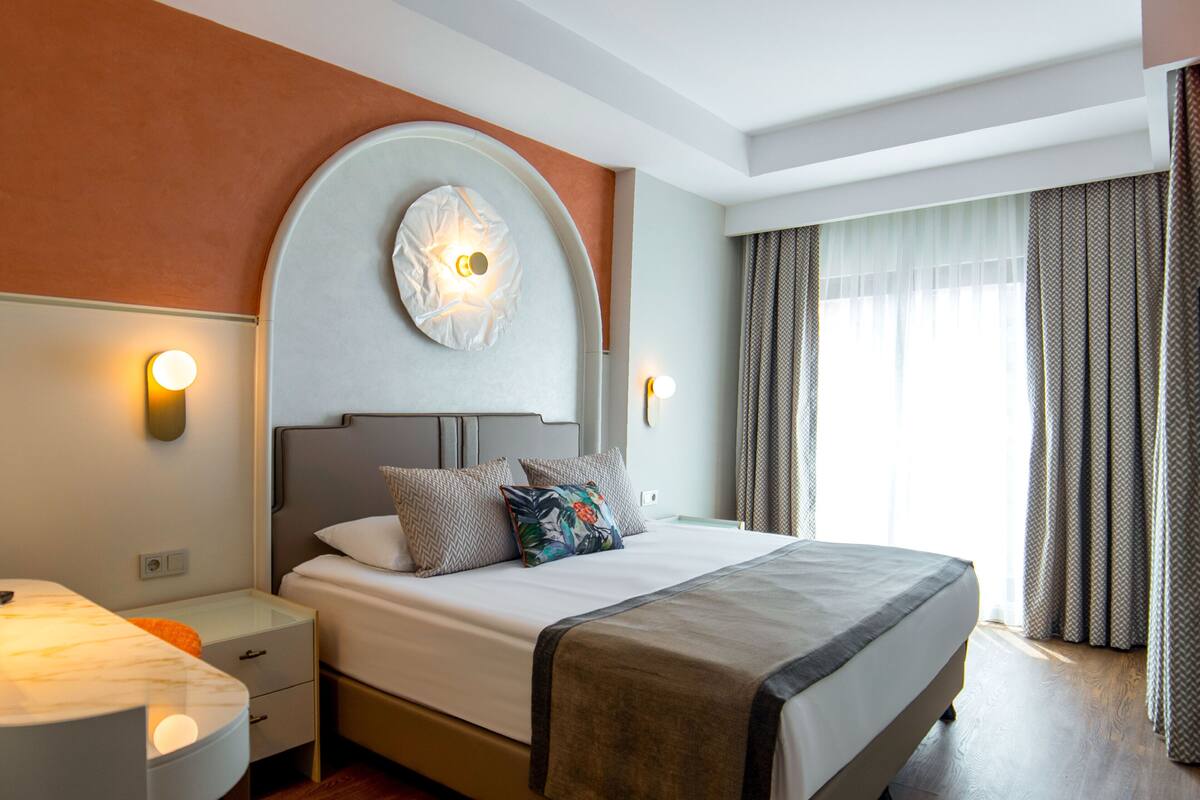 Quarto de hotel com uma cama grande, uma mesa de cabeceira redonda, um criado-mudo e um aplique na parede.