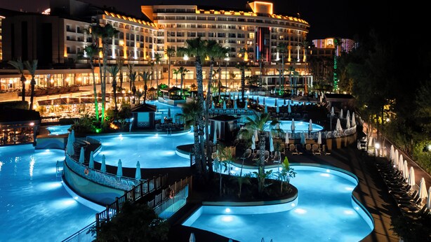 Een hotel met meerdere zwembaden, palmbomen en een goed verlichte gevel 's nachts.