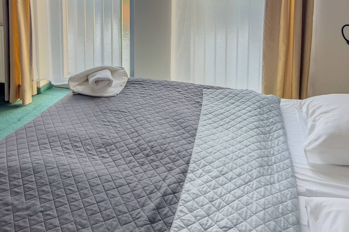 A hotel room with a bed, a rolled towel, and a view of buildings through sheer curtains.