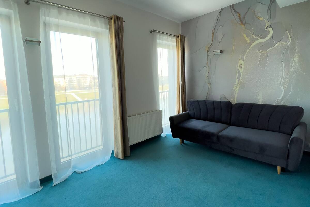 A spacious living room with a dark sofa, large windows with sheer curtains, a radiator, and a wall with a tree-like mural.