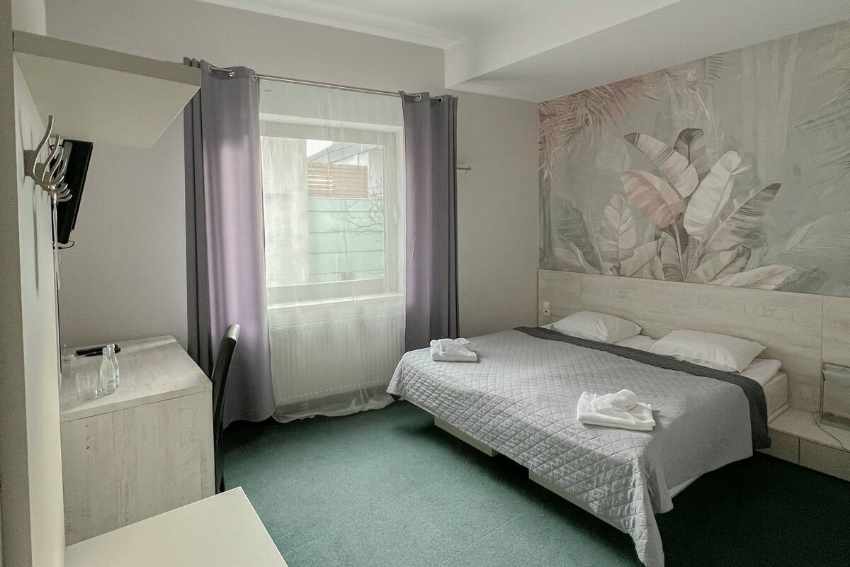 A hotel room with a bed, a desk, a chair, a window with curtains, and a wall with a leaf pattern.
