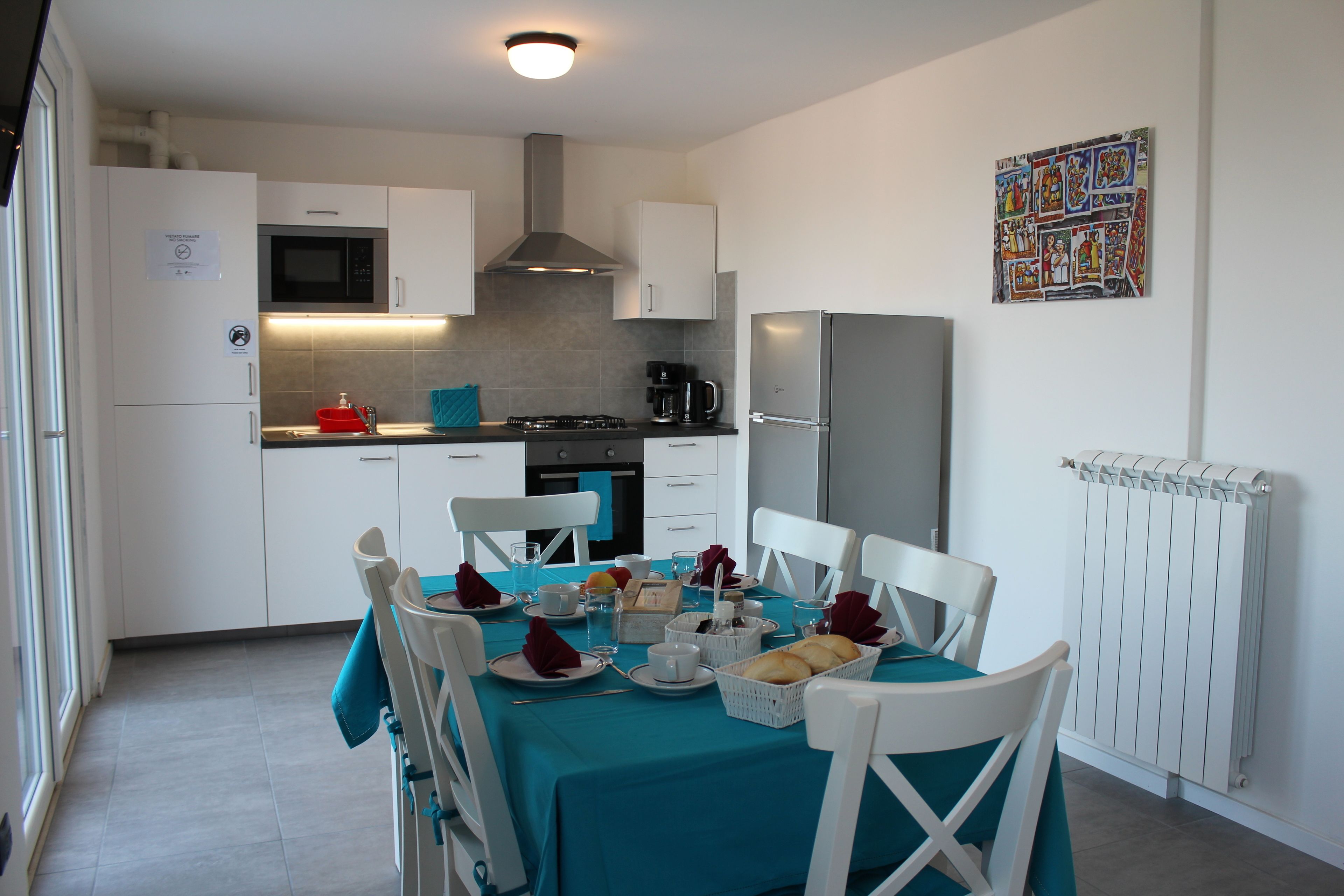 Cocina moderna con armarios blancos, microondas y estufa a gas. Hay una mesa de comedor con sillas blancas y mantel color turquesa.