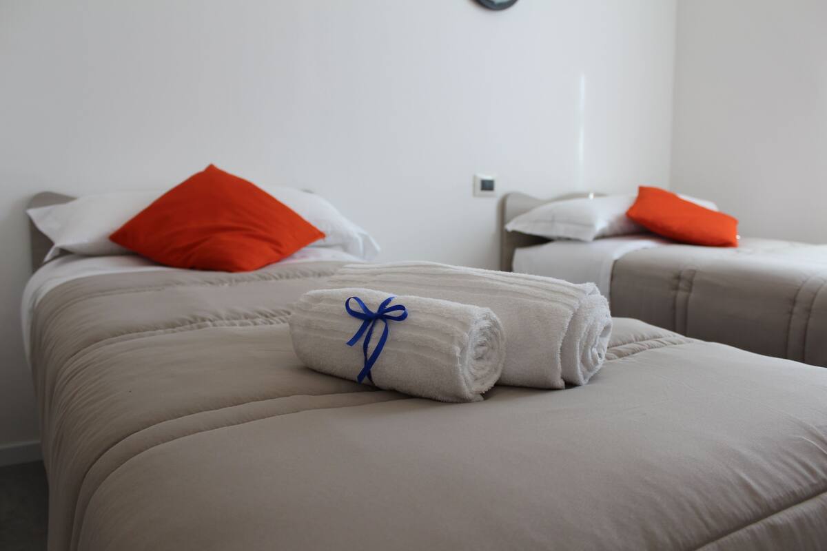 A hotel room with two beds, each with a white pillow and an orange pillow. A rolled white towel with a blue ribbon is placed on the bed in the foreground.