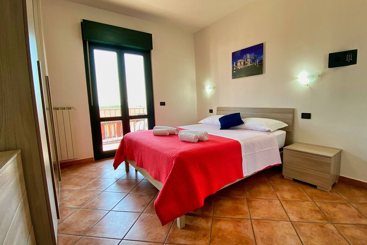 A bedroom with a bed, a nightstand, a wall-mounted light, a painting, and a window with a view.