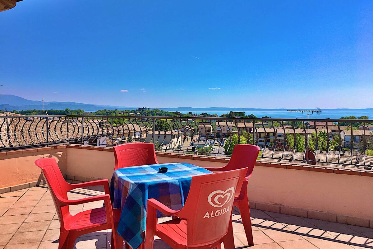 A terrace with a table and chairs, a view of a town and the sea, and a clear sky.