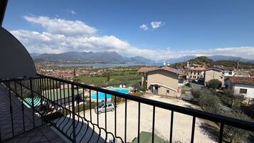 A balcony with a view of a swimming pool, a building with a red roof, and mountains in the distance.