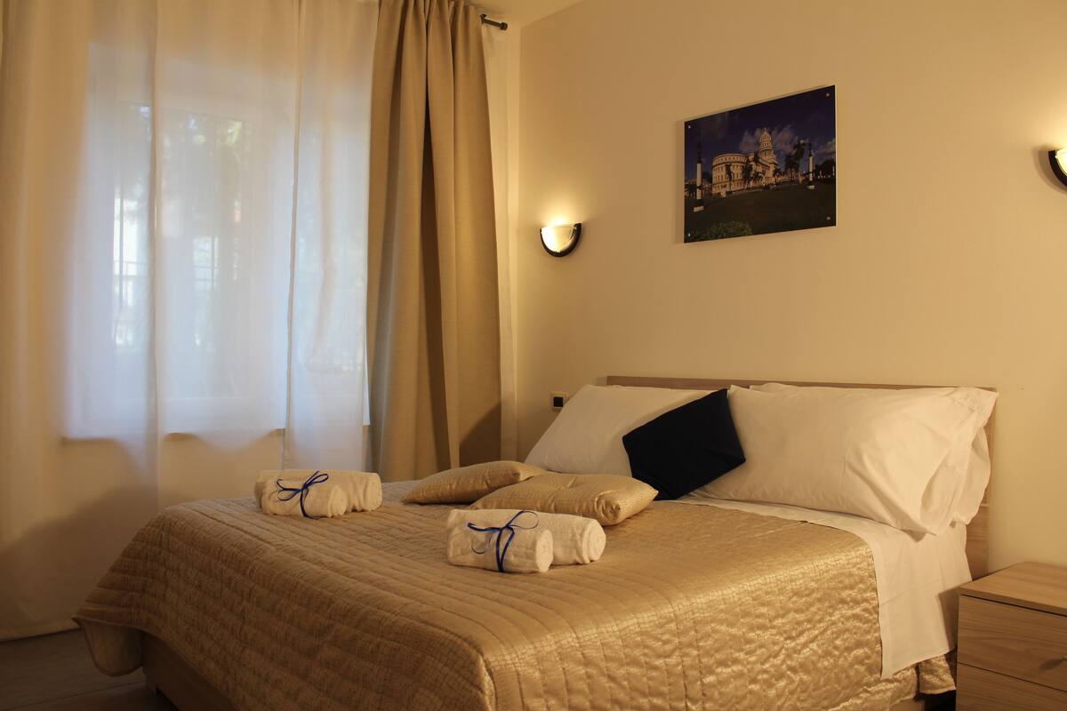 A hotel room with a bed, a nightstand, a wall-mounted light, and a window with curtains.
