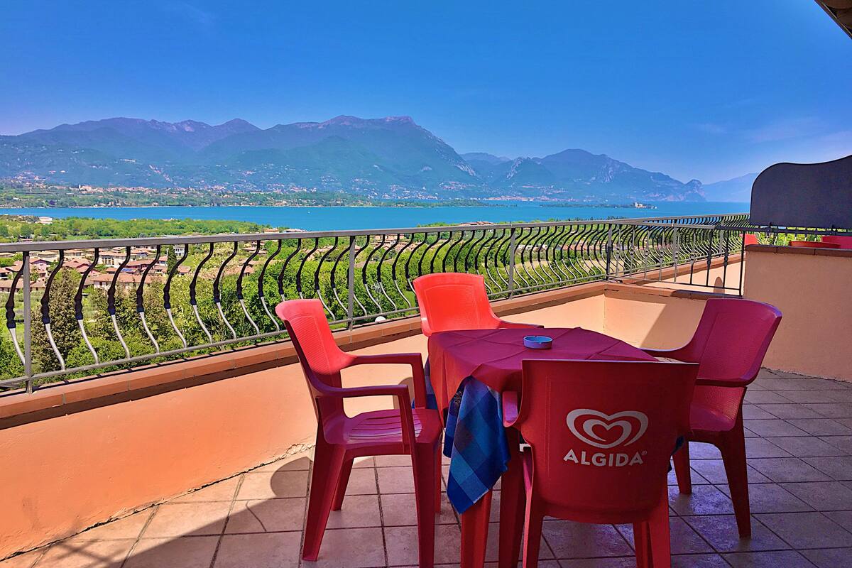 A balcony with red chairs and a table, overlooking a mountainous landscape and a body of water.