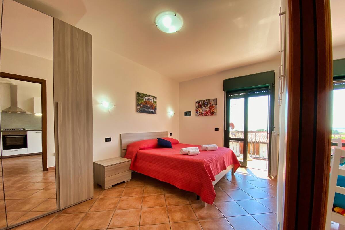 A bedroom with a bed, a nightstand, a mirror, a door to a balcony, and a kitchen visible through an open door.