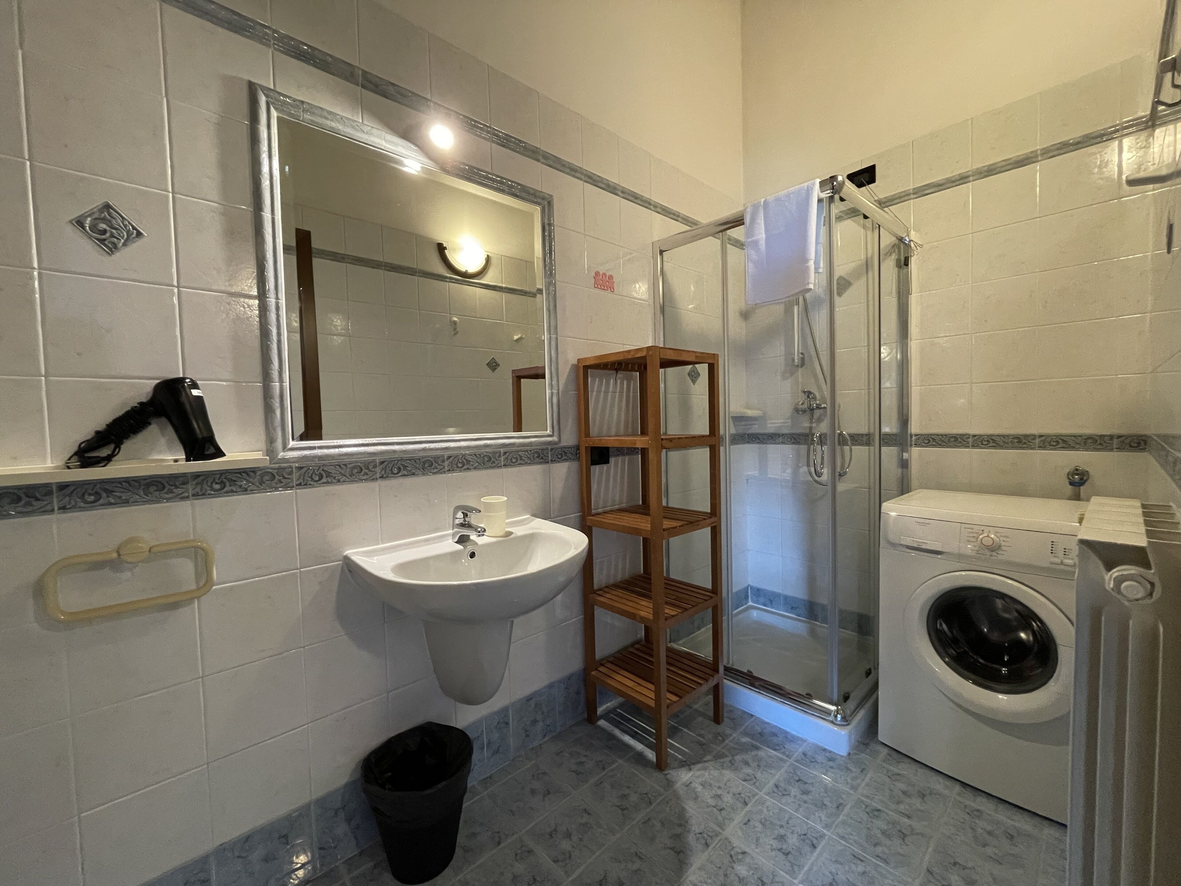 Casa de banho com lavatório, espelho, secador de cabelo, prateleira, chuveiro e máquina de lavar roupa.