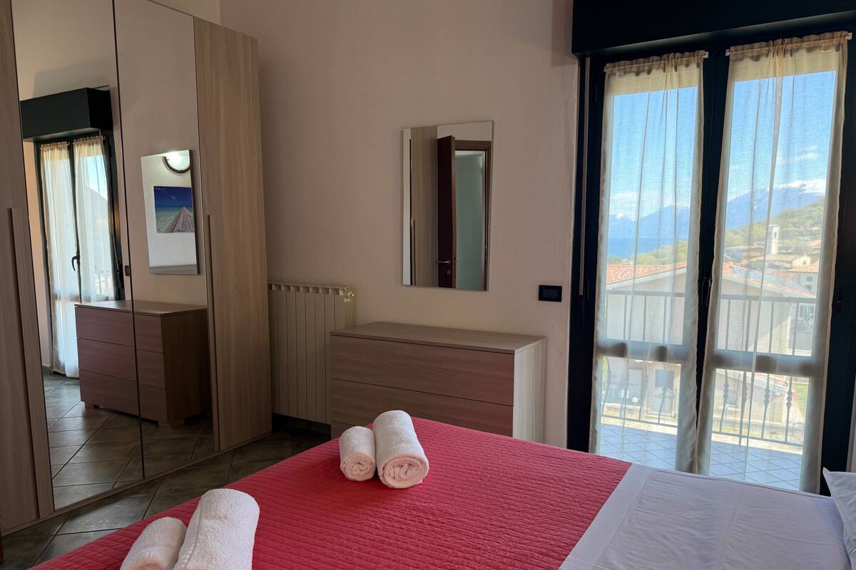 A hotel room with a bed, a red quilted bedspread, a white bedspread, a wooden dresser, a mirror, and a window with a view of buildings and mountains.
