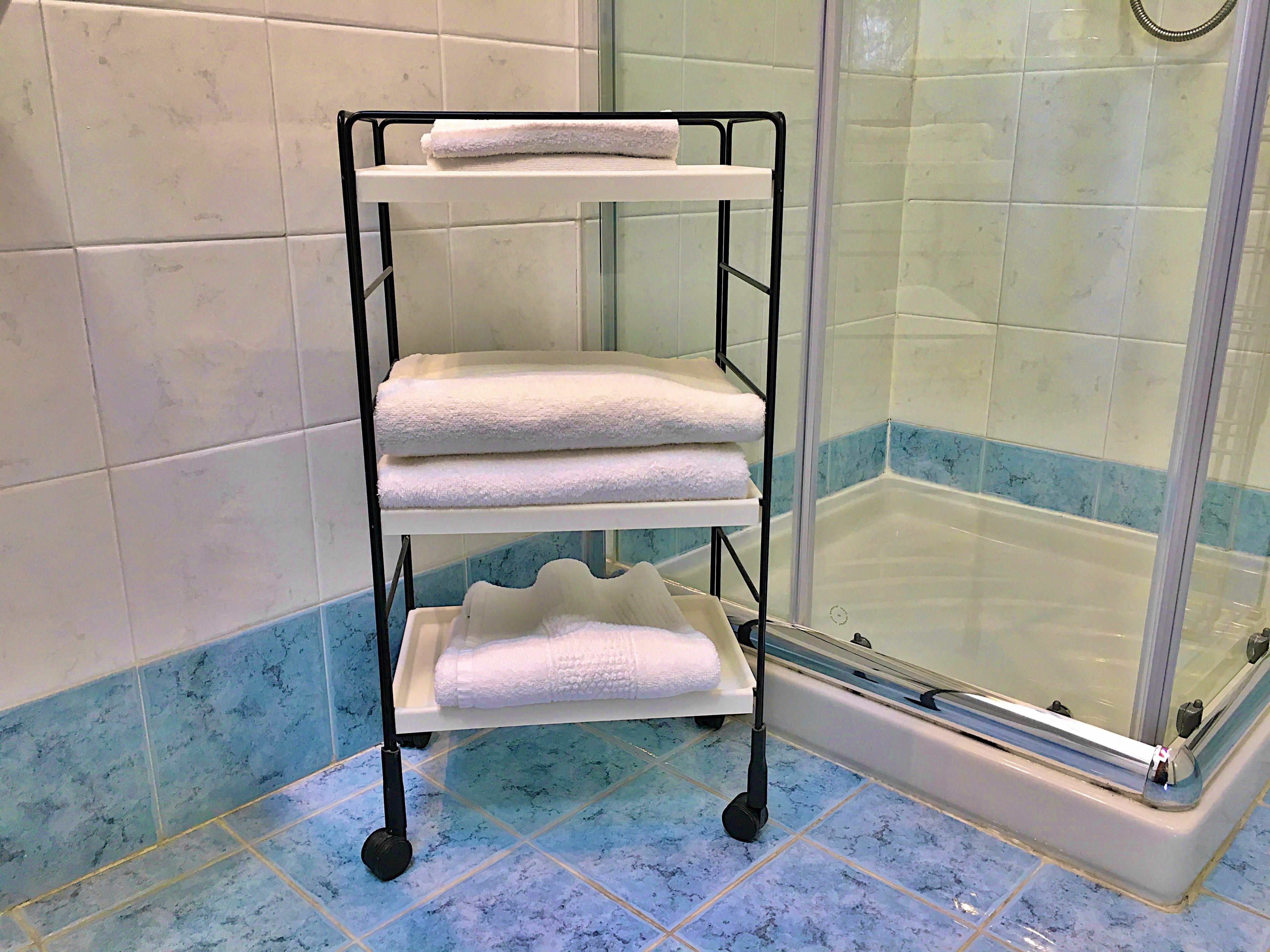 Un bagno con pavimento piastrellato in bianco, zona doccia con porta in vetro e ripiano in metallo nero con asciugamani bianchi.