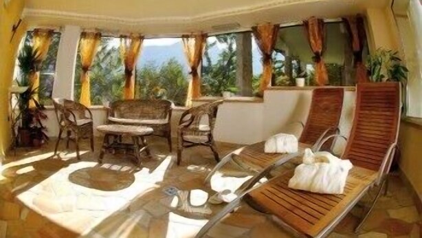 A sunlit room with a table and chairs, a lounge chair, and a window with curtains. - AlpHotel Taller (Dimaro Folgarida)