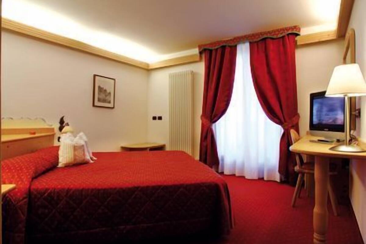 Et hotelværelse med en seng, et skrivebord med lampe, et fjernsyn og et vindue med røde gardiner.