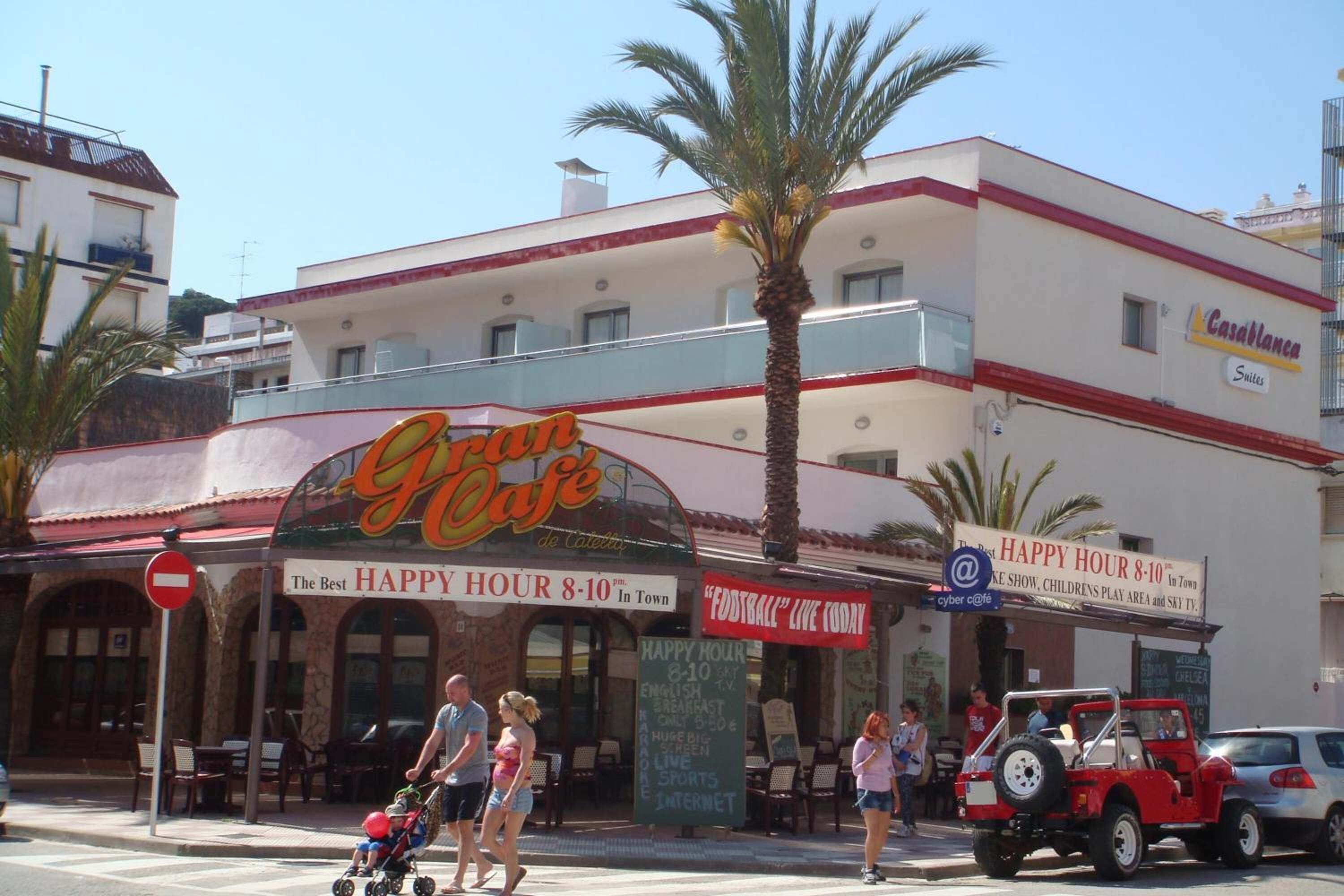 Un café con terraza, un cartel de Happy Hour y un vehículo rojo estacionado afuera.